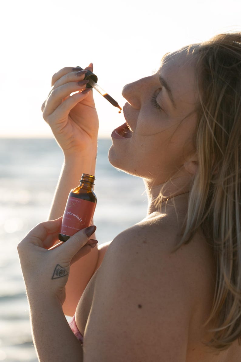 May include: A woman holding a small amber glass bottle and dropper, with the label "Aphrodisia". The woman is tilting her head back and opening her mouth, as a liquid drips from the dropper. The background is a blurred view of the ocean.