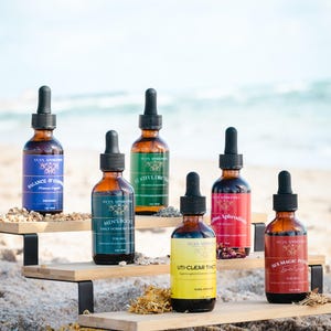May include: A display of brown glass dropper bottles with black caps, each labeled with text and colorful labels. The bottles are arranged on a wooden shelf on a sandy beach, with the ocean in the background. The labels include "Balance & Control", "Men's Boost", and "Sex Magic Potion".