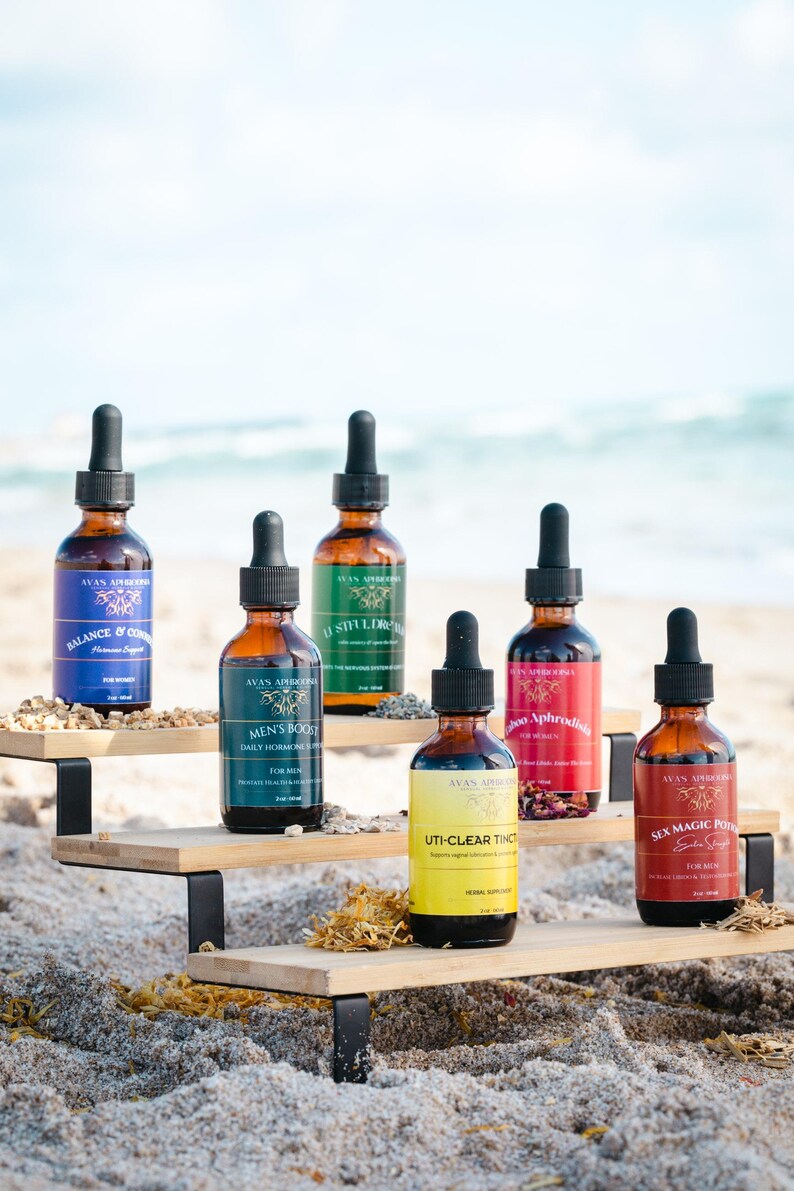 May include: A display of brown glass dropper bottles with black caps, each labeled with text and colorful labels. The bottles are arranged on a wooden shelf on a sandy beach, with the ocean in the background. The labels include "Balance & Control", "Men's Boost", and "Sex Magic Potion".
