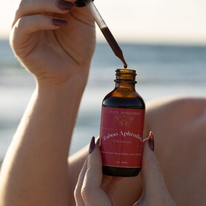 May include: A brown glass bottle with a dropper is held in front of a blurred ocean background. The bottle has a red label with the text "Taboo Aphrodisia FOR WOMEN". The dropper is dispensing a dark liquid. The bottle contains 2 fl oz (60 ml).