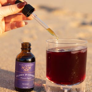 May include: A brown glass bottle with a purple label reading "Balance & Connect Hormone Support" sits next to a glass of red liquid. A dropper dispenses a golden liquid into the glass. The image is set on a sandy beach.