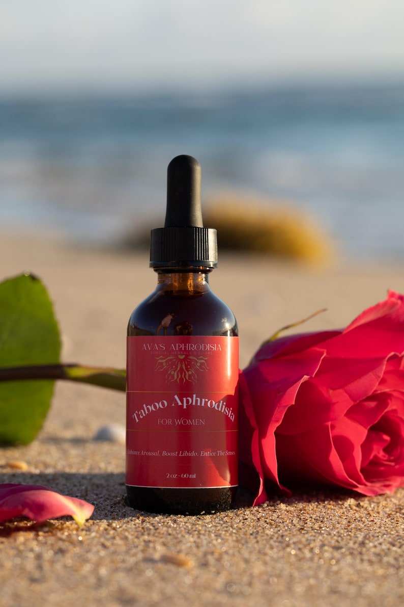 May include: A brown glass bottle with a black dropper, labeled "Taboo Aphrodisia FOR WOMEN" sits on sand next to a red rose. The bottle contains 2 oz (60 ml) of product.