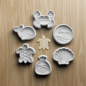 May include: A collection of white plastic cookie cutters in various sea-themed shapes, including a crab, whale, turtle, seashell, wave, and a bag with a fish. The cutters are arranged on a light brown wooden surface.