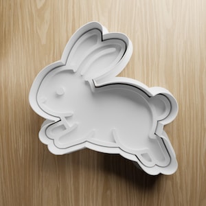 May include: White plastic bunny-shaped cookie cutter. The cutter has a three-dimensional design with a detailed outline of a rabbit in a running pose. The cutter is on a light brown wooden surface.
