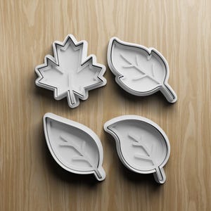 Puede incluir: Cuatro cortadores de galletas blancos con forma de hoja. El juego incluye una hoja de arce y otros tres diseños de hojas. Cada cortador tiene un contorno negro y está sobre un fondo de vetas de madera clara.