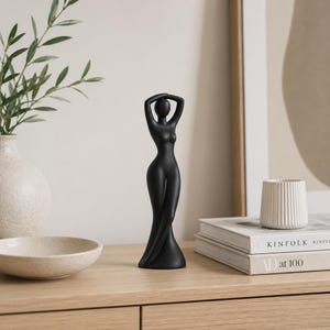Puede incluir: Una escultura negra minimalista de una figura femenina con los brazos levantados sobre la cabeza. La estatua tiene una forma fluida, similar a un vestido. Se exhibe sobre una superficie de madera con otros elementos decorativos.