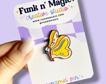 May include: A yellow and cream-colored butterfly enamel pin with a cartoon design. The pin is on a white card with the text "Funk n' Magic creative studio" and "enamel pin". The background is a purple and white checkered pattern.