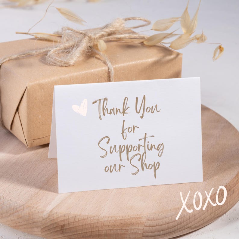 Peut inclure: Une carte blanche avec le texte "Thank You for Supporting our Shop" en lettres brunes et un c&oelig;ur rose. La carte est pos&eacute;e sur une surface en bois avec le texte "XOXO" &eacute;crit en blanc.