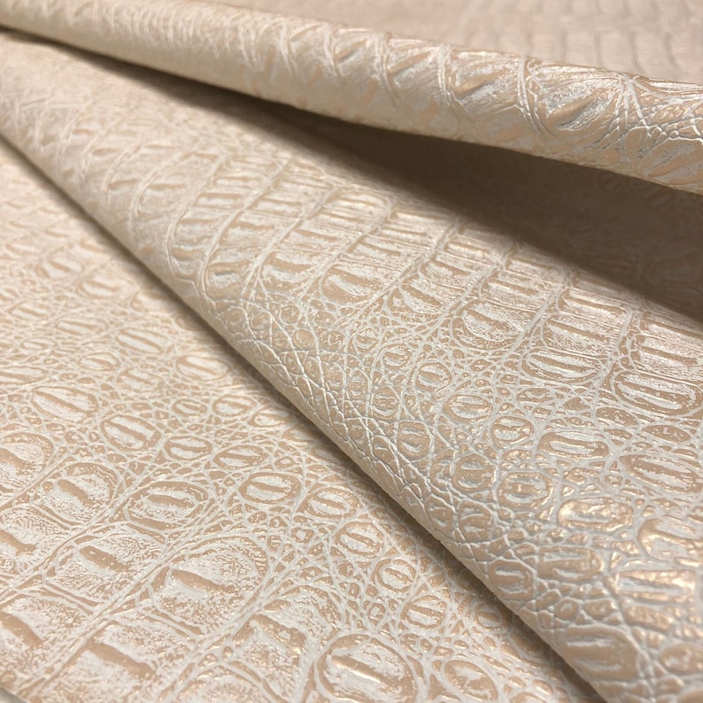 K&ouml;nnte beinhalten: Nahaufnahme eines beigefarbenen, gepr&auml;gten Kunstkrokolederstoffs mit einem subtilen Schimmer. Der Stoff ist in der Mitte gefaltet und zeigt die Textur und das Muster.