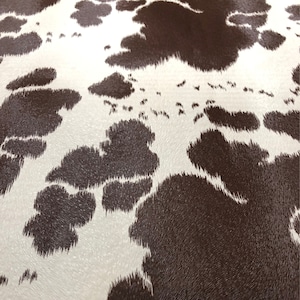 May include: Close-up of a cowhide pattern. The design features irregular dark brown patches against a white background. The texture appears soft and slightly raised, creating a realistic animal hide appearance.