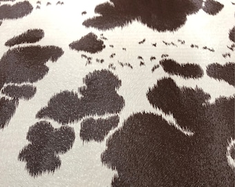 Könnte beinhalten: Nahaufnahme eines Kuhfellmusters. Das Design zeigt unregelmäßige dunkelbraune Flecken auf weißem Hintergrund. Die Textur wirkt weich und leicht erhaben, wodurch ein realistisches Tierfell-Aussehen entsteht.