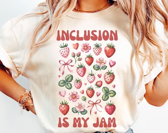 May include: Cream-colored t-shirt with the words "INCLUSION IS MY JAM" in red lettering. The shirt features a pattern of strawberries, flowers, and bows in shades of red and pink.
