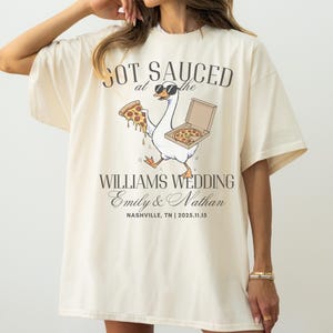 May include: Cream-colored oversized t-shirt with a graphic featuring a goose wearing sunglasses and carrying pizza. Text on the shirt reads "GOT SAUCED at the WILLIAMS WEDDING Emily & Nathan NASHVILLE, TN | 2025.11.15."