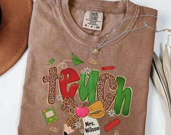 May include: A brown Comfort Colors t-shirt with the word "teach" in a colorful design. The design includes a cross, apple, pencil, and the name "Mrs. Wilson". A silver necklace is draped across the shirt.