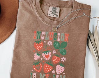 May include: Brown t-shirt with the words "INCLUSION IS MY JAM" in red, green, and pink, along with strawberry graphics. The shirt has a crew neck and short sleeves. A silver necklace with a pendant is draped across the shirt.