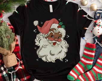 May include: Black t-shirt featuring a Santa Claus graphic with a red hat, white beard, and a smiling face. The Santa graphic has a dark skin tone. The shirt is surrounded by Christmas decorations, including socks and lights.