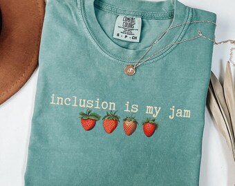 May include: A teal Comfort Colors t-shirt with the phrase "inclusion is my jam" and four strawberry illustrations. A silver necklace with a pendant is draped across the shirt. A brown hat is visible in the upper left corner.