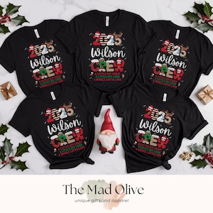 May include: Five black t-shirts with the text "2025 Wilson Crew" in a festive design. The shirts have a crew neck and short sleeves. The text includes reindeer and holiday colors. The Mad Olive logo is at the bottom.