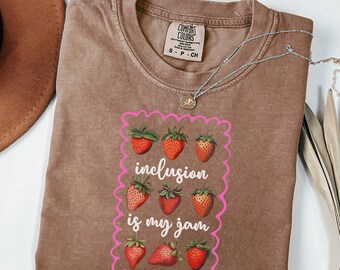 May include: A brown Comfort Colors t-shirt with a pink framed graphic that says "inclusion is my jam" with illustrations of strawberries. A silver necklace with a pendant is on the shirt.