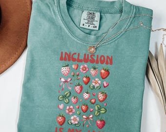 May include: A seafoam green t-shirt with the words "INCLUSION IS MY JAM" in red. The shirt features a strawberry and floral design. A silver necklace with a pendant is draped across the shirt.