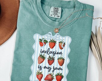 May include: A seafoam green t-shirt with a graphic of strawberries and the phrase "inclusion is my jam." The design is framed by a blue and white striped border. A silver necklace with a pendant is draped across the shirt.