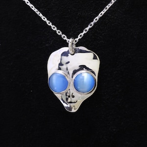 Könnte beinhalten: Ein silberfarbener Anhänger in Form eines Alien-Kopfes, der an einer passenden Kette hängt. Der Anhänger hat eine polierte, reflektierende Oberfläche mit zwei markanten, runden, hellblauen Cabochon-Steinen als Augen und einem kleinen Nasendetail.