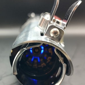 May include: A silver lightsaber hilt with a blue light emitting from the end. The hilt has a detailed design with a series of small, circular indentations.