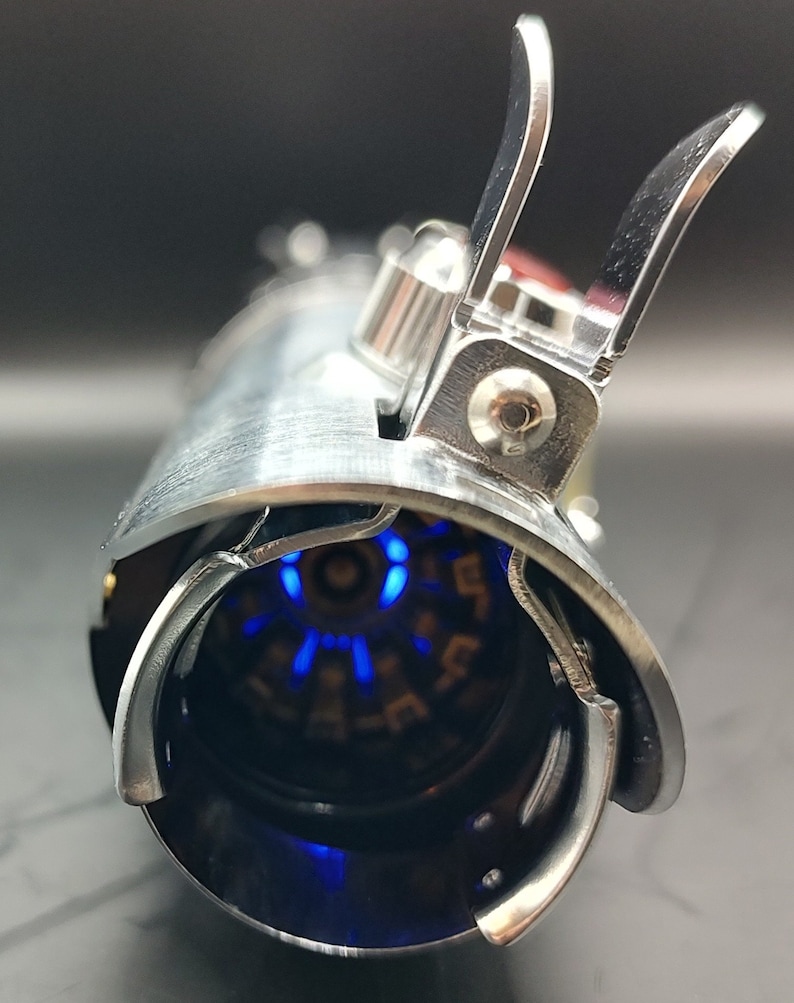 May include: A silver lightsaber hilt with a blue light emitting from the end. The hilt has a detailed design with a series of small, circular indentations.