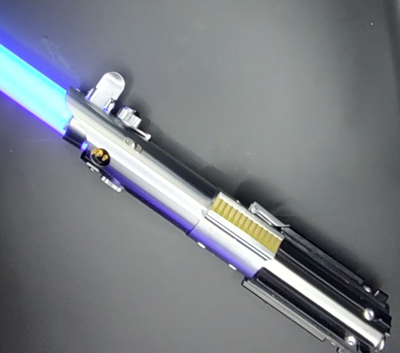 May include: A silver and black lightsaber with a blue blade. The lightsaber has a silver handle with a black grip and a gold accent.