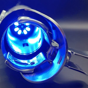 May include: A close-up of a silver sewing machine bobbin case with a blue light shining through the opening. The bobbin case has a lever on the side and a small knob on the top.