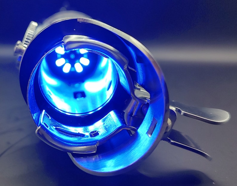 May include: A close-up of a silver sewing machine bobbin case with a blue light shining through the opening. The bobbin case has a lever on the side and a small knob on the top.