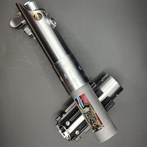 May include: A disassembled lightsaber with a silver body and a black and silver handle. The lightsaber has a circuit board visible inside. The lightsaber is labeled "RAFLLE".