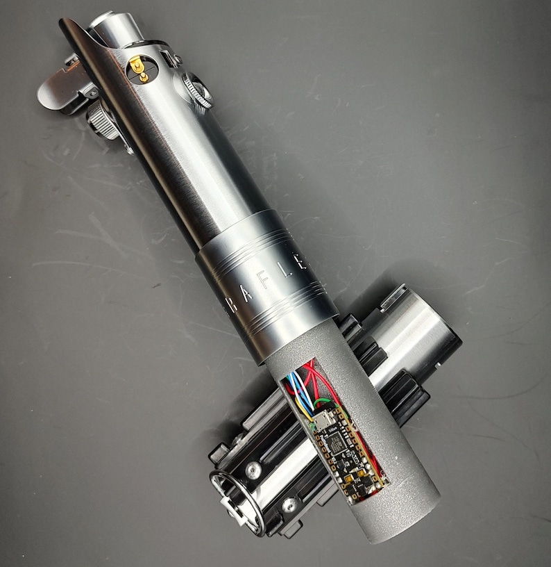 May include: A disassembled lightsaber with a silver body and a black and silver handle. The lightsaber has a circuit board visible inside. The lightsaber is labeled "RAFLLE".