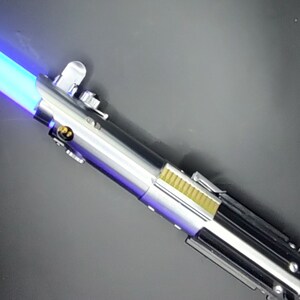 May include: A silver and black lightsaber with a blue blade. The lightsaber has a silver handle with a black grip and a gold accent.