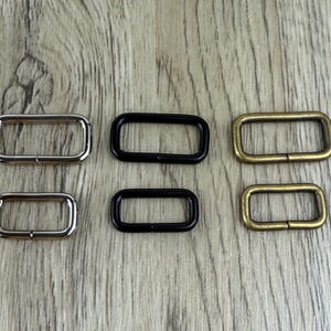 May include: A selection of rectangular metal buckles in silver, black, and brass finishes. The buckles are arranged in a group of six, with two of each colour. The hardware is designed for use in bags, belts, or other crafting projects.
