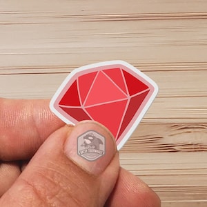 May include: A red gemstone sticker with a white outline. The sticker is held in a hand.