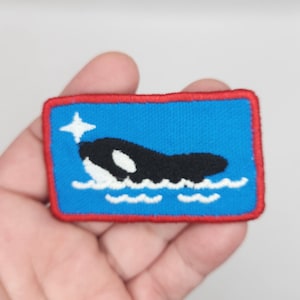 May include: A blue embroidered patch with a red border featuring a white and black orca swimming in blue water with white waves.