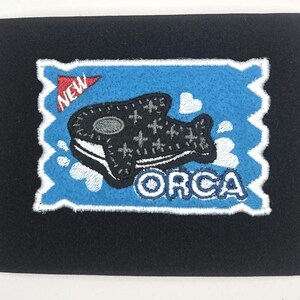May include: A black patch with a blue background featuring a stylized orca whale design. The patch has a white border and the word "ORCA" in white lettering. A red banner with the word "NEW" is in the upper left corner.