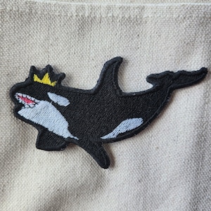 May include: A black and white embroidered patch of a killer whale wearing a yellow crown. The whale is facing left and has a white patch on its belly.