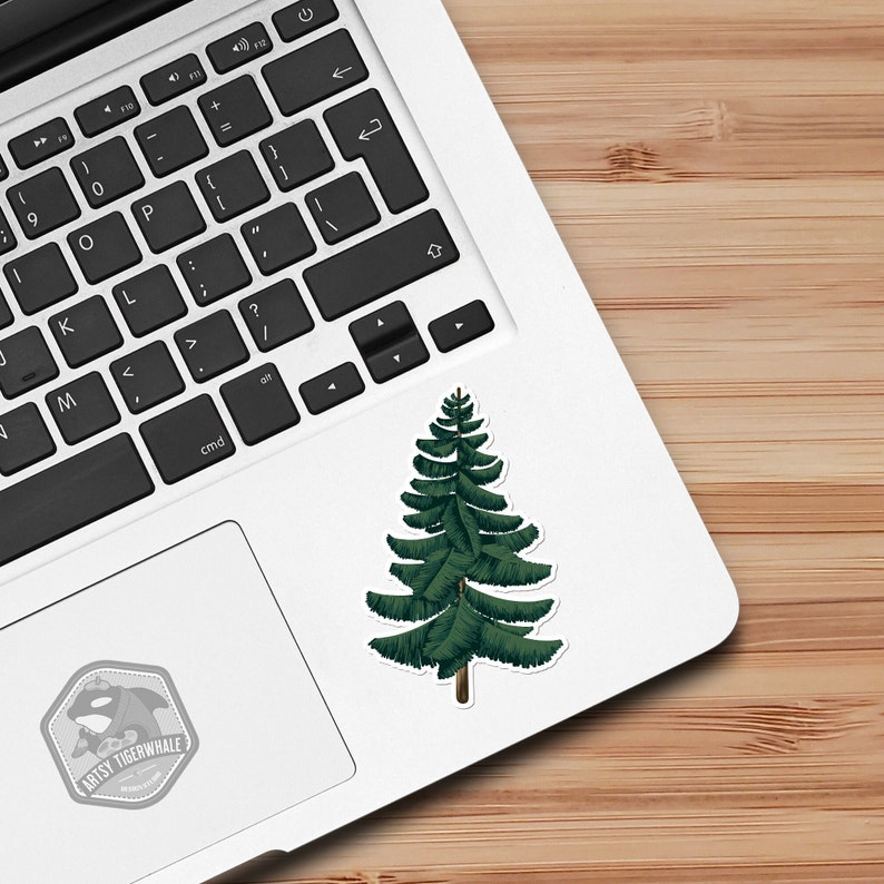 Peut inclure: Un autocollant en forme de pin vert sur un ordinateur portable blanc. L'autocollant est une simple illustration d'un pin avec un tronc brun.
