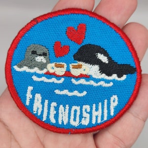 May include: A blue and red embroidered patch with a seal and an orca holding coffee cups. Two red hearts float above them. The text "FRIENDSHIP" is embroidered in white on the blue background.