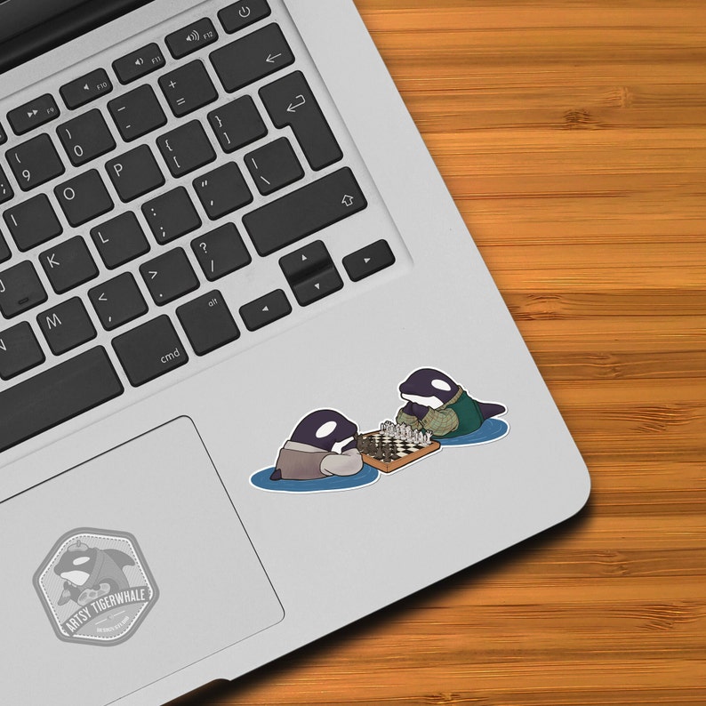 May include: A sticker featuring two cartoon orcas playing chess on a chessboard. The orcas are wearing clothes and are sitting on a blue background. The sticker is on a silver laptop.