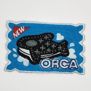 May include: A blue and white embroidered patch with a black and white orca design. The orca is holding a cookie in its mouth. The patch says "NEW" and "ORCA" in white text.