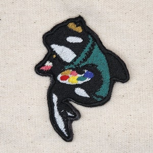 May include: Embroidered iron-on patch featuring a black and white orca holding a colorful paint palette. The orca is wearing a green and black hooded robe.