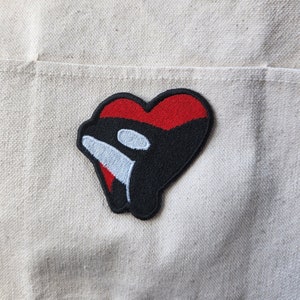 May include: Embroidered patch featuring a black orca whale design within a red heart shape. The orca has a white eye and a white stripe. The patch is on a beige fabric background. This patch is suitable for jackets, bags, or other fabric items.