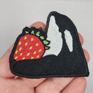 May include: Embroidered patch featuring a black and white orca whale design, with a red strawberry in its mouth. The strawberry has yellow seeds and green leaves. The patch has a black border.