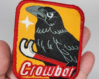 May include: A black crow with a white star above it, holding a red and white crowbar. The crowbar is labeled "Crowbar" in red and white lettering on a yellow background. The patch is surrounded by a red border.