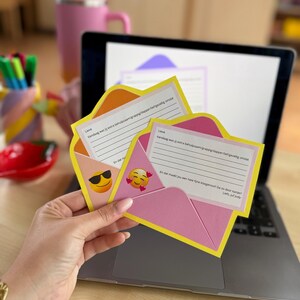 Op de afbeelding: Een roze en gele papieren envelop met een handgeschreven briefje en emoji-stickers. De envelop wordt voor een laptop gehouden. Een roze beker met een rietje en een pennenhouder staan op de achtergrond.