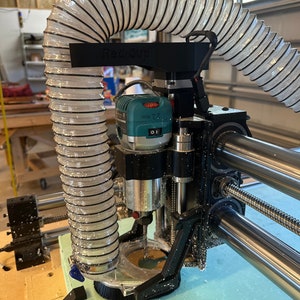 May include: A close-up of a CNC machine with a router bit cutting into a piece of foam. A clear plastic dust collection hose is attached to the router and is connected to a dust collection system. The machine is labeled "RediCup".