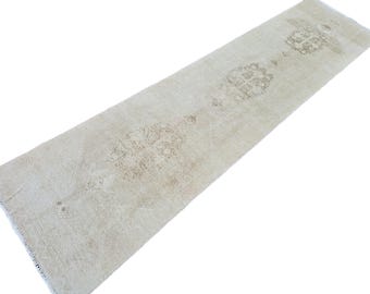 May include: Cream-colored runner rug with a distressed, vintage look. The rug features a repeating pattern of faded floral medallions in shades of beige and brown. The rug has a fringed edge and is ideal for hallways.